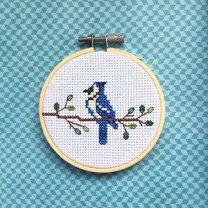 May include: A cross-stitch embroidery of a blue jay perched on a branch with green leaves, set in a wooden hoop. The background is a light blue geometric pattern.