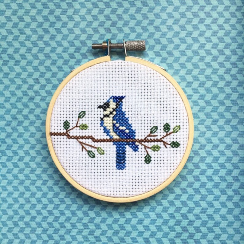 May include: A cross-stitch embroidery of a blue jay perched on a branch with green leaves, set in a wooden hoop. The background is a light blue geometric pattern.