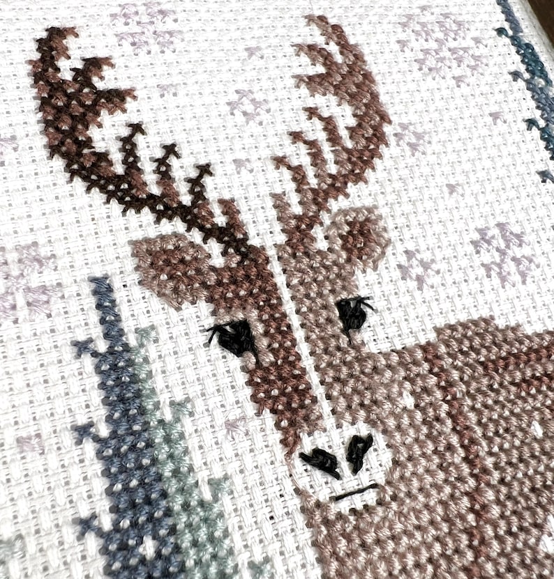 May include: A cross-stitch design featuring a deer with brown antlers and a textured, patterned body. The artwork is set against a white background with subtle, light-colored cross-stitch details, creating a rustic, handcrafted aesthetic.