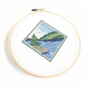 May include: A cross-stitch embroidery hoop with a diamond-shaped design of a lake scene. The scene features a green hill, a blue lake with white dots, a green tree, and two white birds flying in the sky.