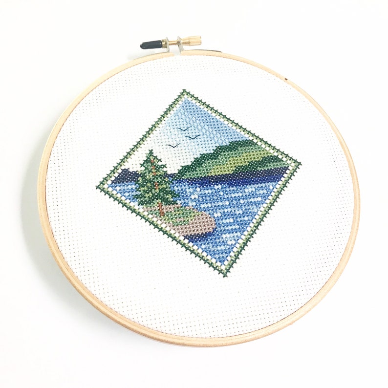 May include: A cross-stitch embroidery hoop with a diamond-shaped design of a lake scene. The scene features a green hill, a blue lake with white dots, a green tree, and two white birds flying in the sky.
