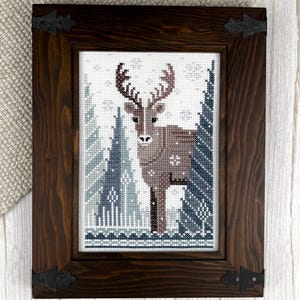 May include: Cross-stitch artwork of a deer in a winter forest scene, framed in a dark brown wooden frame with decorative corner brackets. The artwork features a brown deer with large antlers, surrounded by blue and white trees and snowflakes.