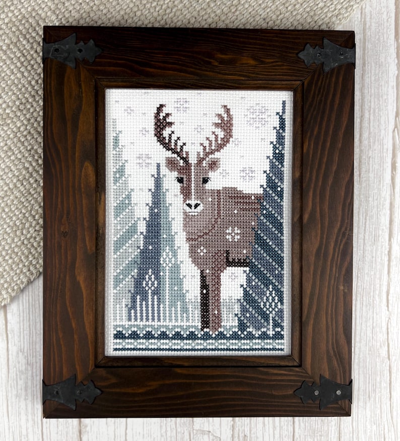 May include: Cross-stitch artwork of a deer in a winter forest scene, framed in a dark brown wooden frame with decorative corner brackets. The artwork features a brown deer with large antlers, surrounded by blue and white trees and snowflakes.