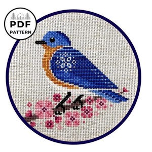 May include: A cross-stitch design featuring a bluebird on a branch with pink flowers, framed by a navy circle. The bird is blue and orange. The text "PDF PATTERN" is in a black circle.