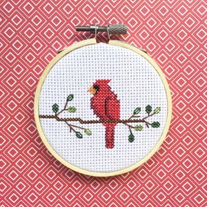 May include: A cross-stitch embroidery hoop featuring a red cardinal bird perched on a brown branch with green leaves. The bird is set against a white background, framed by a wooden hoop. The background is a red pattern.
