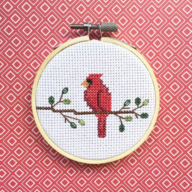 May include: A cross-stitch embroidery hoop featuring a red cardinal bird perched on a brown branch with green leaves. The bird is set against a white background, framed by a wooden hoop. The background is a red pattern.