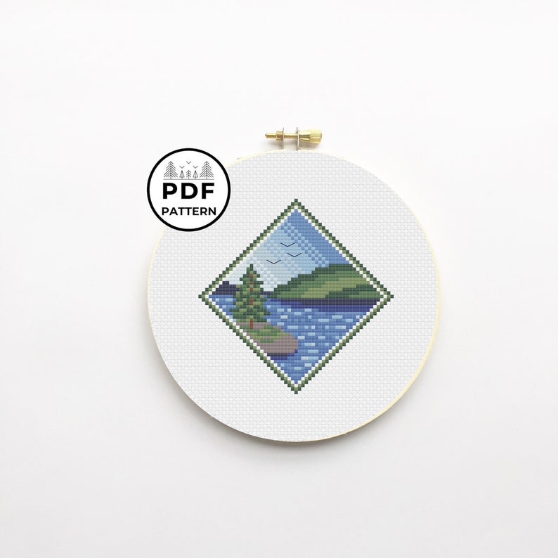 May include: A diamond-shaped cross-stitch pattern featuring a lake scene with a green tree, blue water, and a green hill in the background. There are three birds flying in the sky above the lake.