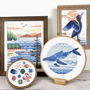 Pu&ograve; includere: Quattro ricami a punto croce incorniciati che rappresentano una balena blu, un uccello blu, un paesaggio marino e una collezione di conchiglie. I disegni sono esposti su una superficie di legno bianca.