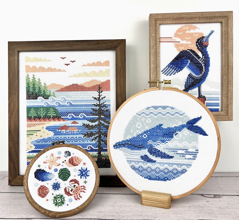 Pu&ograve; includere: Quattro ricami a punto croce incorniciati che rappresentano una balena blu, un uccello blu, un paesaggio marino e una collezione di conchiglie. I disegni sono esposti su una superficie di legno bianca.