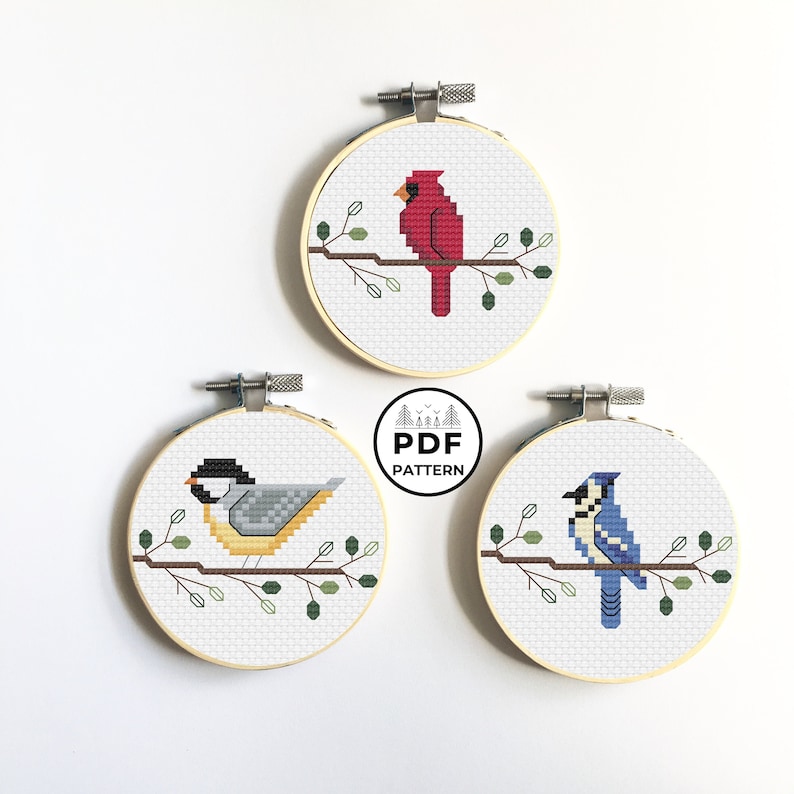 May include: Three cross-stitch embroidery hoops featuring birds: a red cardinal, a gray and yellow bird, and a blue jay. Each bird is perched on a brown branch with green leaves, set against a white background. The text "PDF PATTERN" is in the center.