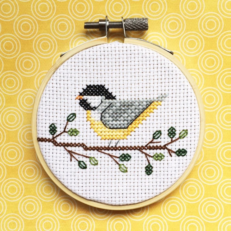 May include: A cross-stitch embroidery of a bird perched on a branch with leaves. The bird has a black head, gray wings, and a yellow breast. The embroidery is in a wooden hoop, set against a yellow background.