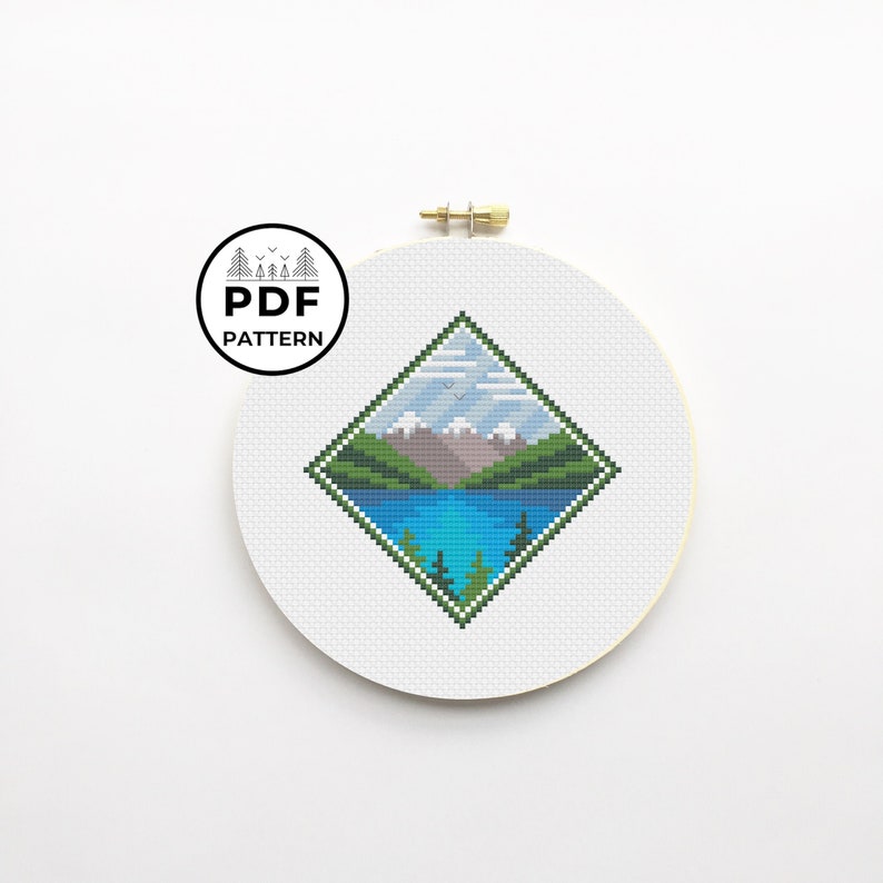 May include: A cross-stitch pattern of a mountain landscape with a lake, trees, and a blue sky. The pattern is in a diamond shape and is framed in a white embroidery hoop.