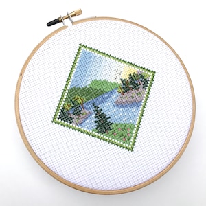 Puede incluir: Un aro de bordado blanco con un marco de madera. El aro contiene un dise&ntilde;o de punto de cruz de una escena de lago con &aacute;rboles verdes, un lago azul y un cielo azul claro.