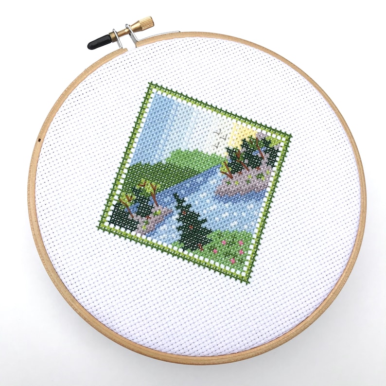 Puede incluir: Un aro de bordado blanco con un marco de madera. El aro contiene un dise&ntilde;o de punto de cruz de una escena de lago con &aacute;rboles verdes, un lago azul y un cielo azul claro.