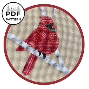 May include: A cross-stitch pattern featuring a bright red cardinal bird with a black face mask perched on a white branch against a beige fabric background. The circular image includes a circular logo in the top left corner with the text PDF PATTERN.