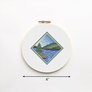 May include: A 6-inch round embroidery hoop with a cross-stitch design of a lake scene. The design features a green hill, a blue lake, a small green tree, and three white birds flying in the sky.