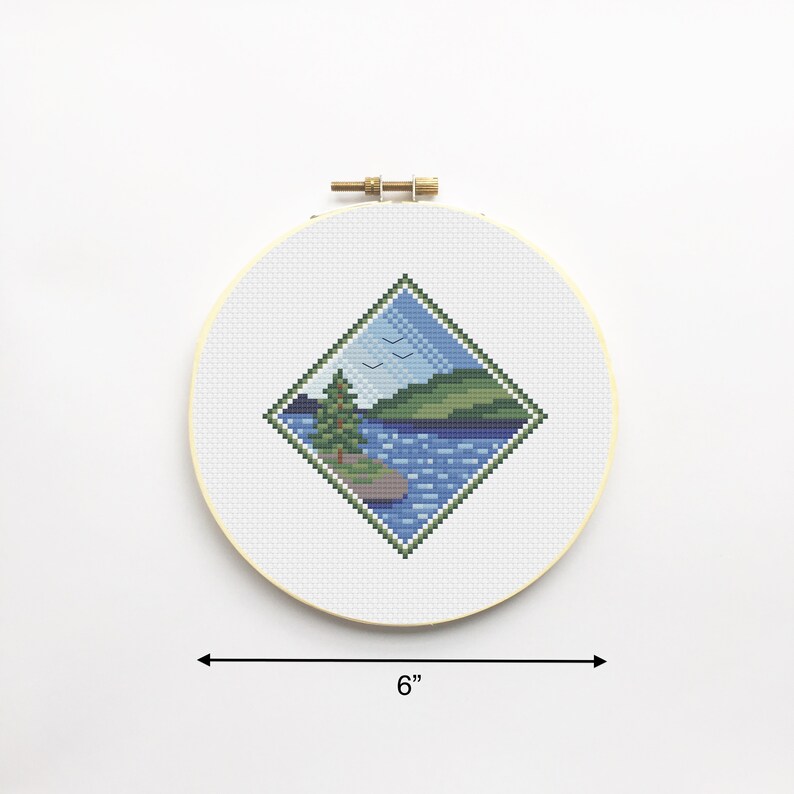 May include: A 6-inch round embroidery hoop with a cross-stitch design of a lake scene. The design features a green hill, a blue lake, a small green tree, and three white birds flying in the sky.