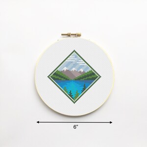 May include: A 6-inch round embroidery hoop with a white background and a diamond-shaped cross-stitch design of a mountain lake scene. The scene features a blue lake, green mountains, and a light blue sky with white clouds.