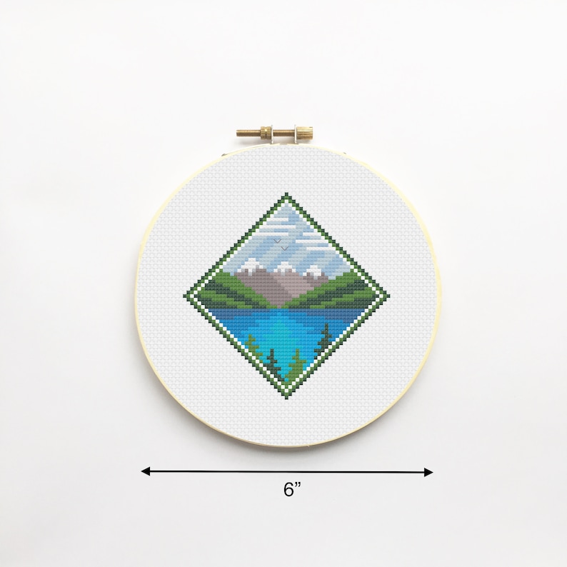 May include: A 6-inch round embroidery hoop with a white background and a diamond-shaped cross-stitch design of a mountain lake scene. The scene features a blue lake, green mountains, and a light blue sky with white clouds.