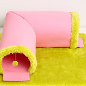 May include: A pink cat tunnel with lime green edging and a matching green plush mat. A small green ball hangs inside the tunnel entrance. The cat bed is designed in a modern style.