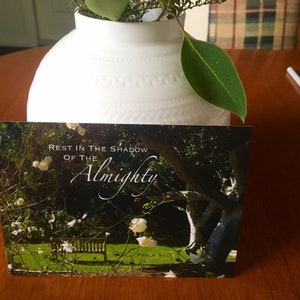 May include: A sympathy card with a white vase and green leaves in the background. The card features a photo of a tree with white flowers and a bench in a garden. The text on the card reads "Rest in the shadow of the Almighty, Psalm 91:1".