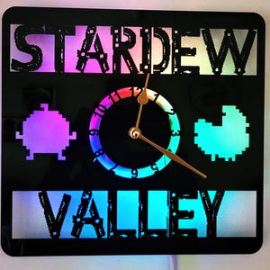 Könnte beinhalten: Schwarze quadratische Wanduhr mit den ausgeschnittenen Worten "STARDEW VALLEY", beleuchtet mit farbigen LED-Leuchten. Das Zifferblatt hat einen Regenbogenverlauf und goldene Zeiger. Pixelige Bilder von Charakteren befinden sich an den Seiten.