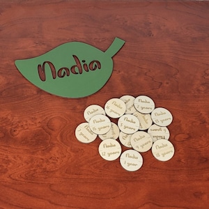 May include: A green leaf-shaped wooden cutout with the name "Nadia" cut out in the center.  There are 18 wooden circles with the name "Nadia" and a number of years written on each circle.