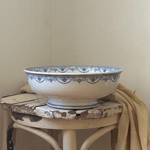 May include: A white ceramic wash basin with a blue floral pattern along the rim, resting on a distressed white wooden stool with three legs. A beige fabric cloth drapes over the side of the stool, which shows signs of chipped paint and wear.