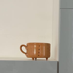 May include: A small, brown ceramic mug with a handle and three small feet. The mug has a cylindrical shape with evenly spaced, small, dark dots around the body. The mug is set against a backdrop of light gray and white surfaces.
