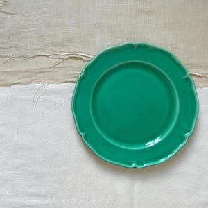 Peut inclure: Une assiette en céramique vert vif avec un bord festonné. L'assiette est posée sur un fond de tissu blanc cassé texturé et un morceau de lin beige. La finition brillante de l'assiette contraste avec les textures naturelles.