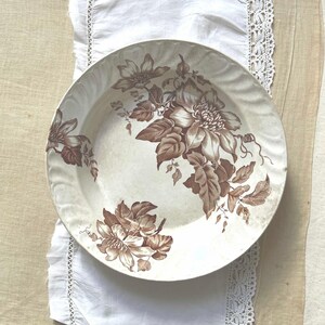 Könnte beinhalten: Ein weißer Keramikteller mit braunem Blumendesign. Der Teller liegt auf einem weißen Tuch mit Spitzenbesatz, auf einer beigefarbenen Leinenoberfläche. Das Blumenmuster zeigt große Blüten und Blätter.