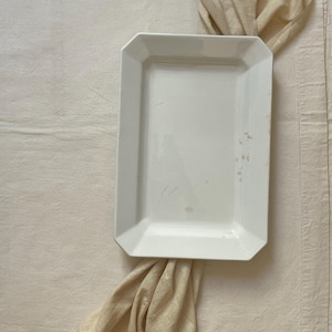 Può includere: Un piatto da portata rettangolare in ceramica bianca con un bordo leggermente rialzato. Il piatto presenta alcune piccole macchie marroni. Un tessuto beige è drappeggiato attorno al piatto su una superficie color crema.