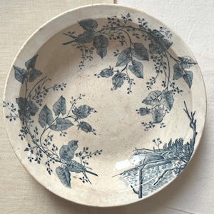 Peut inclure: Assiette ancienne en céramique crème avec un motif floral et paysager bleu. L'assiette présente un motif botanique détaillé de feuilles et de vignes sur le bord, avec une illustration scénique au centre. L'assiette a un aspect légèrement usé.