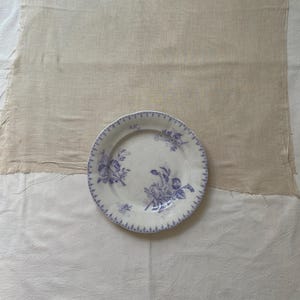 May include: A white ceramic plate with a blue floral design and a scalloped edge. The plate is set on a beige linen cloth over a white cloth, creating a rustic, vintage aesthetic.