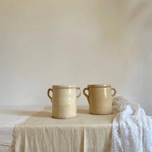 Può includere: Due antichi vasi in ceramica bianco sporco con manici. I vasi hanno una forma cilindrica e un bordo leggermente più scuro. Sono appoggiati su una superficie di lino beige, con un panno di pizzo bianco drappeggiato sul lato destro.