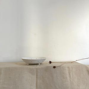 Pode incluir: Uma tigela escorredor de cerâmica branca com pequenos orifícios de drenagem espaçados uniformemente e três pés curtos e arredondados. A tigela tem um design raso com borda larga e está posicionada ao lado de um galho seco e fino.