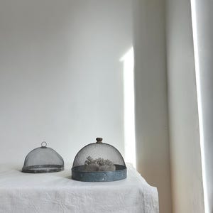 Puede incluir: Dos campanas de malla metálica para alimentos sobre un mantel blanco. Una es en forma de cúpula con un pomo de madera, la otra es una cúpula más pequeña y plana. Las campanas están sobre una mesa con tela de lino blanca.