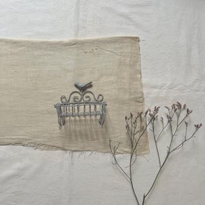 Pode incluir: Um pequeno pássaro de metal prateado empoleirado em um banco decorativo de arame. O banco está sobre uma superfície de tecido bege. À direita da imagem, há um ramo de flores rosa secas.