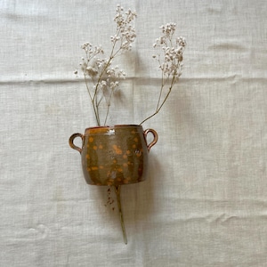 Pode incluir: Um pequeno vaso de cerâmica castanho com duas pegas, cheio de delicadas flores brancas. O vaso tem um acabamento salpicado e vidrado com detalhes em laranja. As flores são altas e esguias, adicionando um toque de natureza ao design rústico.
