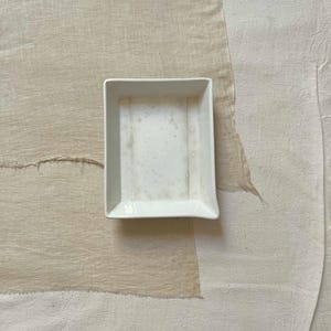 May include: A small, square, white ceramic dish with slightly raised edges. The dish has a simple, minimalist design and appears to be clean. The background is a neutral color, with a textured fabric.