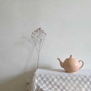 May include: A peach-colored ceramic teapot with a rounded body, spout, and handle sits on a white lace tablecloth. A delicate dried flower stem stands to the left. The background is a plain, off-white wall.