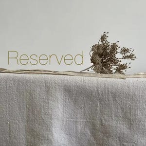 May include: A neutral-toned linen fabric with the word "Reserved" in a gold colour. A small bundle of dried flowers rests on the fabric. The fabric has a textured appearance and is likely used for home decor or textiles.
