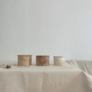 May include: Three small, cylindrical ceramic pots in varying shades of beige and brown are arranged on a cream-coloured linen tablecloth. The pots have a rustic, aged appearance, suggesting a vintage or handcrafted origin. The background is a plain white wall.