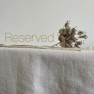 Peut inclure: Un tissu en lin de couleur neutre avec le mot "Reserved" en or. Un petit bouquet de fleurs séchées repose sur le tissu. Le tissu a un aspect texturé et est probablement utilisé pour la décoration intérieure ou les textiles.