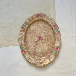 Könnte beinhalten: Eine ovale Servierplatte aus Keramik mit gewelltem Rand, die in der Mitte einen Blumenstrauß aus rosa, gelben und blauen Blüten zeigt, umgeben von einem dichten braunen Blumenmuster. Der Rand ist mit einer Girlande aus bunten Blüten verziert.
