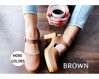 May include: Brown leather clogs with a buckle strap and a wooden heel. The clogs are worn by a person sitting on a wooden floor. The text "MORE COLORS" and "BROWN" are visible in the image.