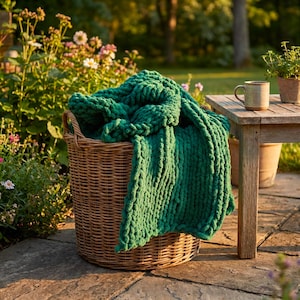 May include: A woven wicker basket containing a thick, emerald green knitted blanket. The basket is on a stone patio. A wooden table with a mug and potted plants is in the background, with flowers and greenery surrounding the scene. The blanket is a vibrant green.