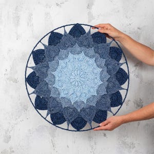 Può includere: Un arazzo da parete rotondo all'uncinetto blu con un design sfumato. Il centro è azzurro, che sfuma in tonalità di blu più scure verso il bordo esterno. Il pezzo è tenuto da mani e ha una cornice blu scuro.