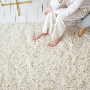 May include: A white fluffy rug with a long pile. The rug is on a white wooden floor.