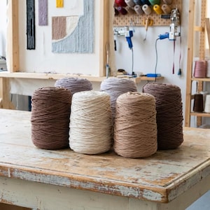 Puede incluir: Seis madejas de hilo de varios colores, incluyendo marrón, beige y gris, están dispuestas sobre una mesa de madera envejecida. El hilo está bien enrollado, lo que sugiere que está listo para proyectos de manualidades. El fondo muestra un estudio de manualidades.